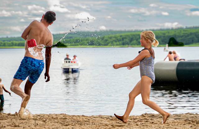 Papa und Tochter laufen über Sandstrand in Badesachen, im Hintergrund Labussee mit Personen auf dem Wasser