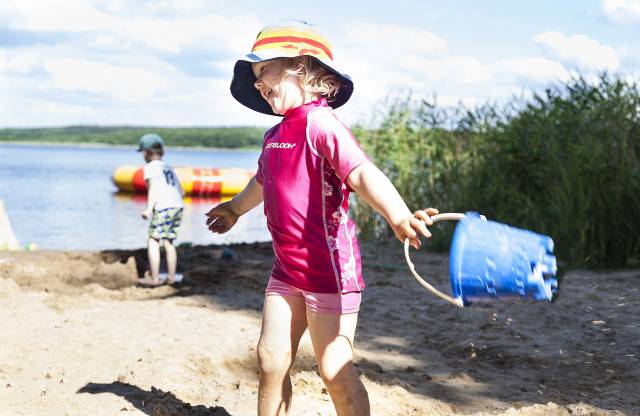 Kleines Mädchen mit Hut und Schwimmkleidung, blauem Eimer in der Hand lacht am Strand