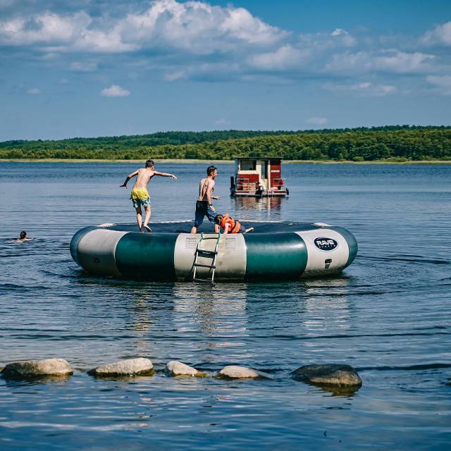 eine Familie springt auf großem Wassertrampolin im Wasser, im Hintergrund ein kleines Hausboot und eine Person schwimmt im Wasser