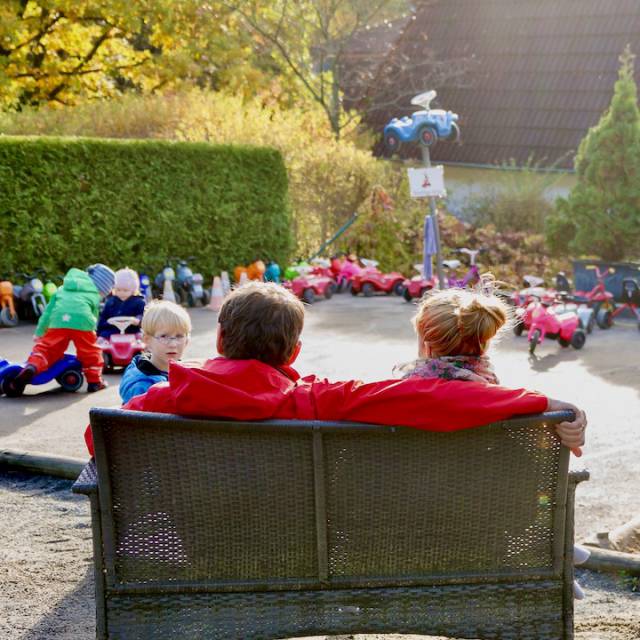 Eltern sitzen in der Sonne und schaue den Kindern beim Bobbycar fahren zu