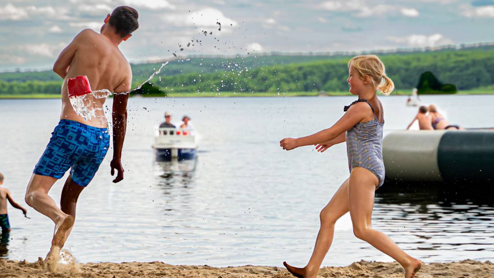 Papa und Tochter laufen über Sandstrand in Badesachen, im Hintergrund Labussee mit Personen auf dem Wasser