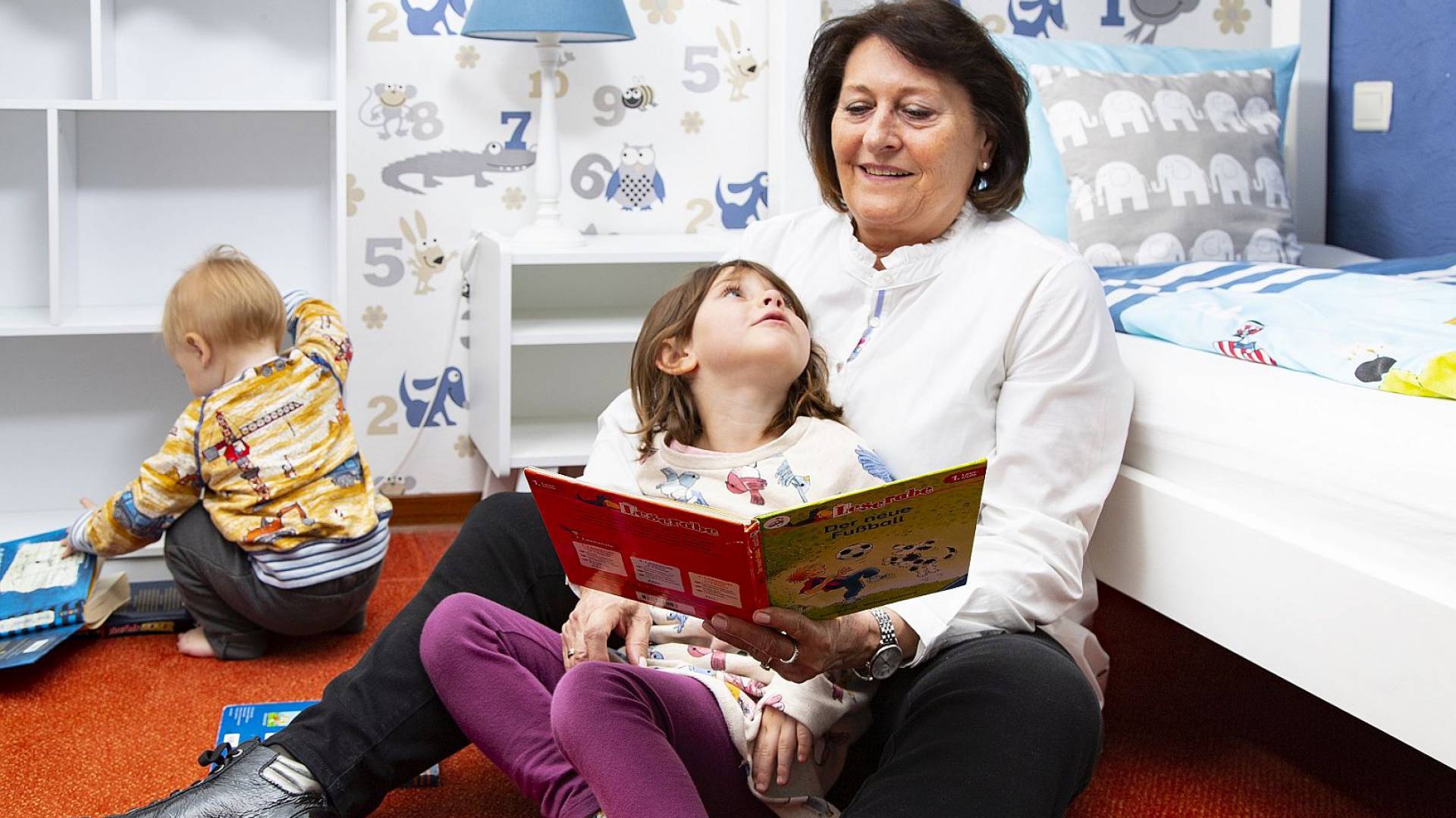  Im Borchards Rookhus liest die Mutter der Enkelin aus einem Buch vor währen die Enkelin im Schoß sitzt und hoch schaut, daneben sitz ein kleiner Junge und blättert im Buch