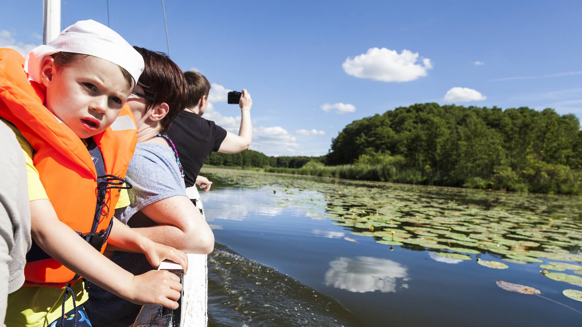 Eine Familie fährt mit dem Boot über die mecklenburgische Seenplatte. Man sieht blauen Himmel und viele Seerosen.